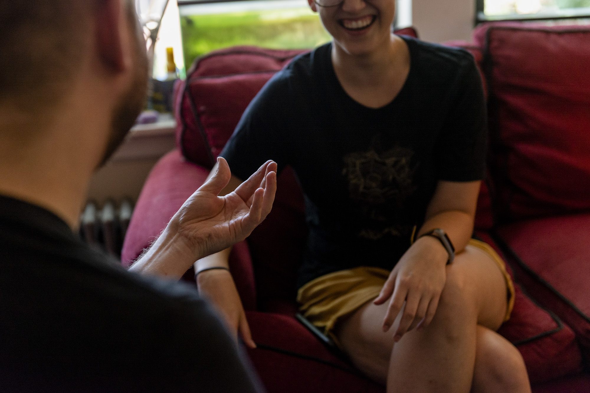 Image: The viewer is looking over the artists shoulder as he makes a hand gesture that resembles a "cupping" motion. Across from the artist is his roommate, their legs are crossed with one arm resting on their left leg. The top half of their face is out of the frame but their bottom half is laughing. Photo by Ryan Edmund Thiel.