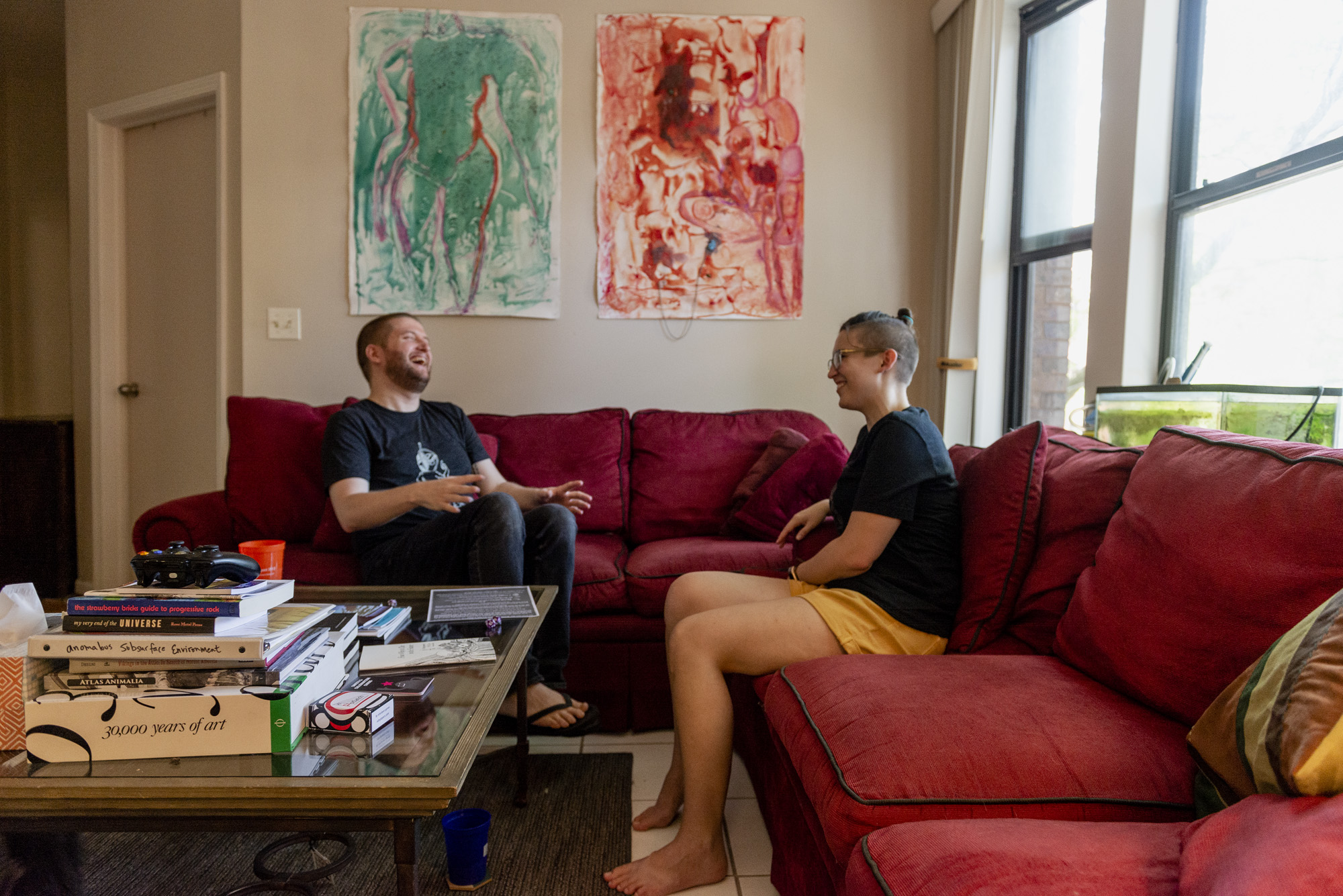 Image: This is a full view of the living room with two red couches. One subject is sitting on the red couch to the right of the frame and the artist is sitting in the middle of the frame on the red couch against the back wall. There is a coffee table in the middle with books and other objects piled on top. Hung on the back wall are two paintings created by Lamar Gayles Jr., one is green/blue and the other is orange/red in hue. The artist is laughing and slightly leaning back and the other subject is also laughing. Photo by Ryan Edmund Thiel