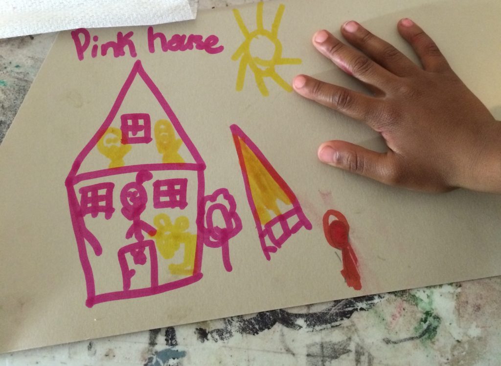 Image: A drawing at Pink House, St. Louis, held down by a child’s hand. The drawing is bright pink and yellow on white paper. The drawing shows a cross-section of the house with people inside on the first and second floors; outside the pink house are the words “Pink house,” a sun, trees, and more. Photo by Patrick Fuller. Courtesy of the artist.