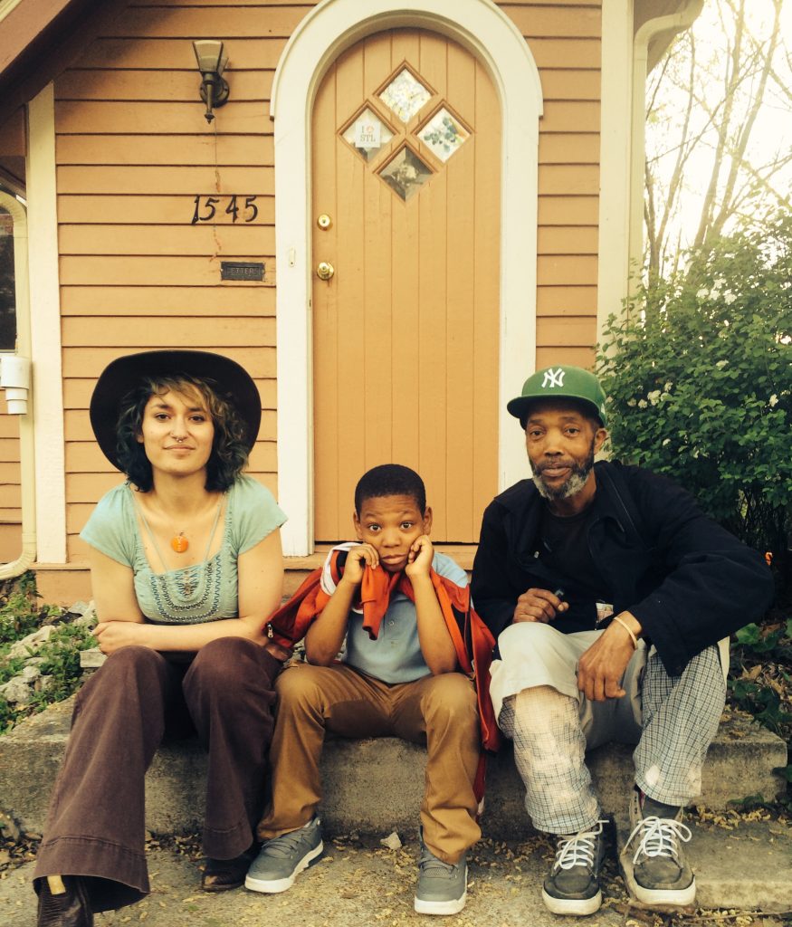 Image: Regina Martinez at Pink House in St. Louis in April 2016, with Javontá Fletcher and Curtis Lomax (left to right). The three sit on Pink House’s concrete front step, posing for the camera. Martinez wears a broad-brimmed hat, a patterned sea-foam green shirt, and brown pants; Fletcher wears a light blue shirt and brown pants, and holds an orange jacket around his shoulders; Lomax wears a black jacket, light shorts over light plaid pants, and a green New York Yankees cap. Martinez and Lomax are adults and Fletcher is a child. Photo by Joy Southerland. Courtesy of the artist.