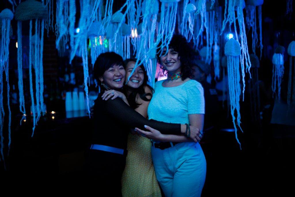 Image: Regina Martinez at “the clothesline” in St. Louis in 2019. Martinez (right) poses with co-founders April Fulstone and Angelina Fasano. They stand together, smiling at the camera with arms around each other, in a dark space lit by bluish light. Hanging from the ceiling and all around them are dozens of crocheted jellyfish made of light-colored yarn. Fulstone wears all black with a blue belt, Fasano wears a yellow and white patterned outfit, and Martinez wears a light top and light pants with a bright, multi-colored choker. Photo by Carly Faye. Courtesy of the artist.