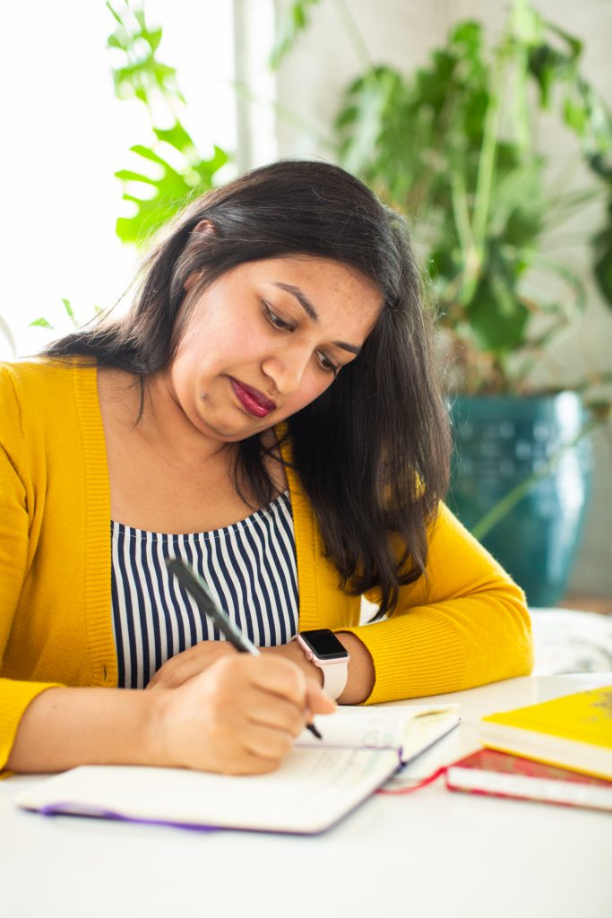 Image: Sharanya Sharma. Sharma sits at a white table, looking down at an open notebook. Sharma holds a pen poised above the paper. Sharma wears a marigold cardigan open over a black and white striped shirt. Behind Sharma is a large plant, and natural light comes through the windows. Photo by Kristie Kahns Photography.