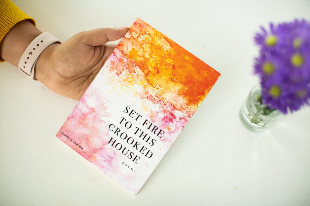 Image: A copy of Sharanya Sharma’s MFA thesis, a poetry collection entitled “Set Fire to This Crooked House.” From off-camera, Sharma’s hand angles the thesis toward the camera. The thesis is bound with a colorful cover, on which orange and pink watercolors fade into each other and into white. The title appears in all capital letters and the words “poems” and “sharanya sharma” in all lower-case. On the white tabletop is a vase of purple and yellow flowers. Photo by Kristie Kahns Photography.