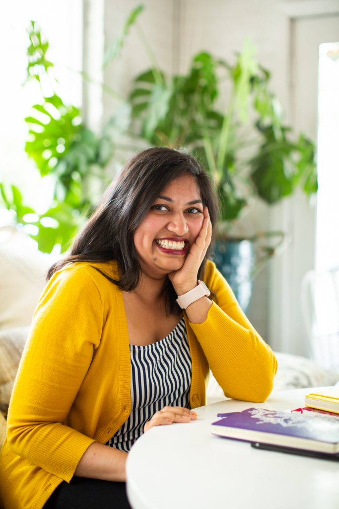 Image: Sharanya Sharma. Sharma sits, leaning on one hand, elbow on a white table next to a few multi-colored notebooks. Sharma wears a marigold cardigan open over a black and white striped shirt and smiles at the camera. Behind Sharma is a large plant, and natural light comes through the windows. Photo by Kristie Kahns Photography.