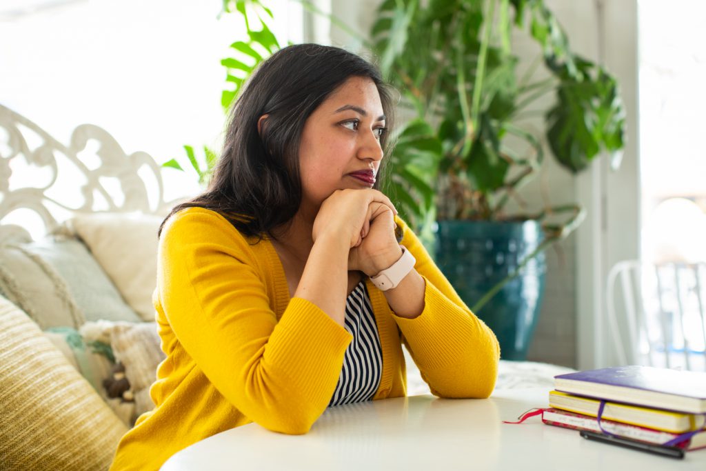 Image: Sharanya Sharma. Sharma sits with hands under chin and elbows on a white table, next to a pile of multi-colored notebooks. Sharma wears a marigold cardigan open over a black and white striped shirt and looks off, out of the frame. Behind Sharanya are several pastel throw pillows and a large plant, and natural light comes through the windows. Photo by Kristie Kahns Photography.