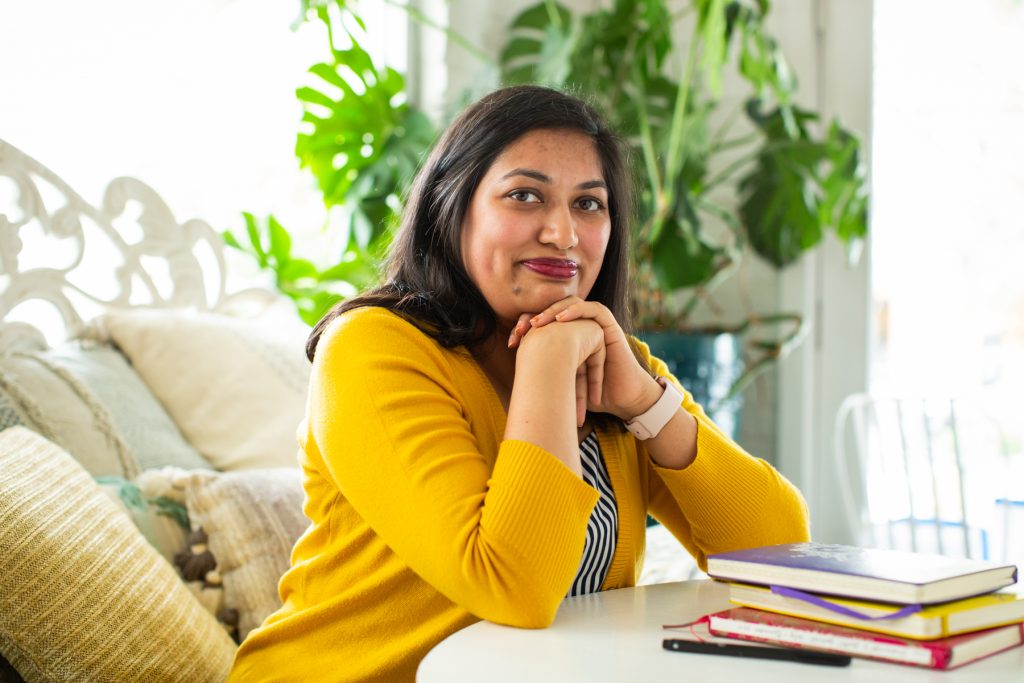 Image: Sharanya Sharma. Sharma sits with hands under chin and elbows on a white table, next to a pile of multi-colored notebooks. Sharma wears a marigold cardigan open over a black and white striped shirt and looks at the camera with a slight smile. Behind Sharma are several pastel throw pillows and a large plant, and natural light comes through the windows. Photo by Kristie Kahns Photography.