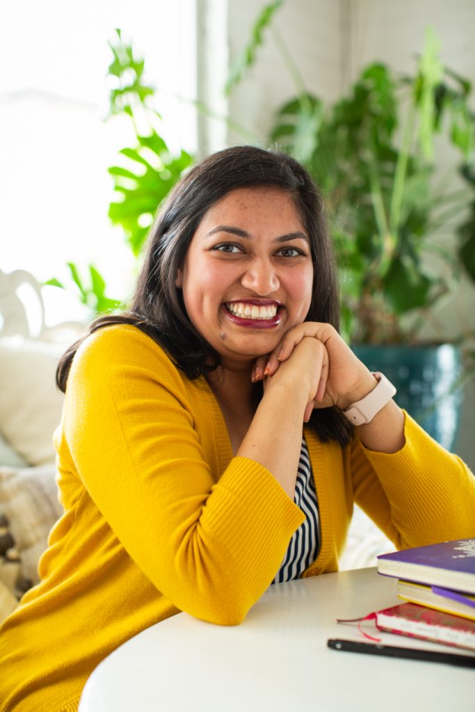 Image: Sharanya Sharma. Sharma sits, leaning on one hand, elbow on a white table next to a few multi-colored notebooks. Sharma wears a marigold cardigan open over a black and white striped shirt and smiles at the camera. Behind Sharma is a large plant, and natural light comes through the windows. Photo by Kristie Kahns Photography.