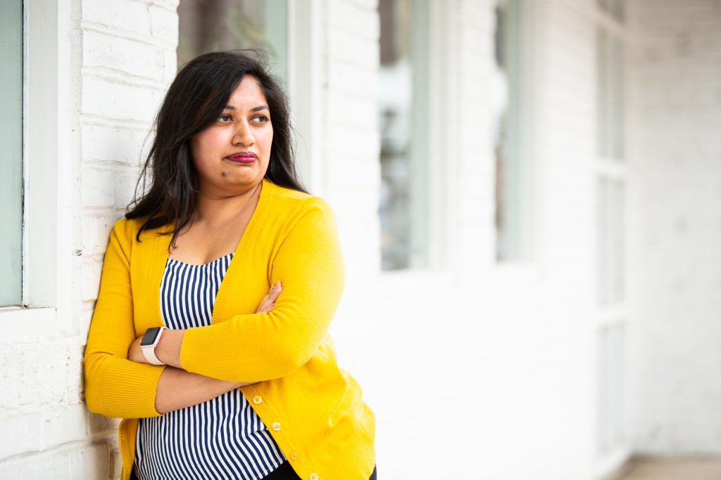 Image: Sharanya Sharma. Sharma leans against the outside of a white brick building, looking off-camera. Sharma wears a marigold cardigan open over a black and white striped shirt, and Sharma’s arms are folded. Photo by Kristie Kahns Photography.