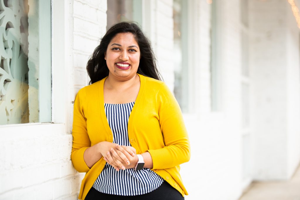 Image: Sharanya Sharma. Sharma leans against the outside of a white brick building, smiling at the camera. Sharma wears a marigold cardigan open over a black and white striped shirt, and Sharma’s hands are folded. Photo by Kristie Kahns Photography.