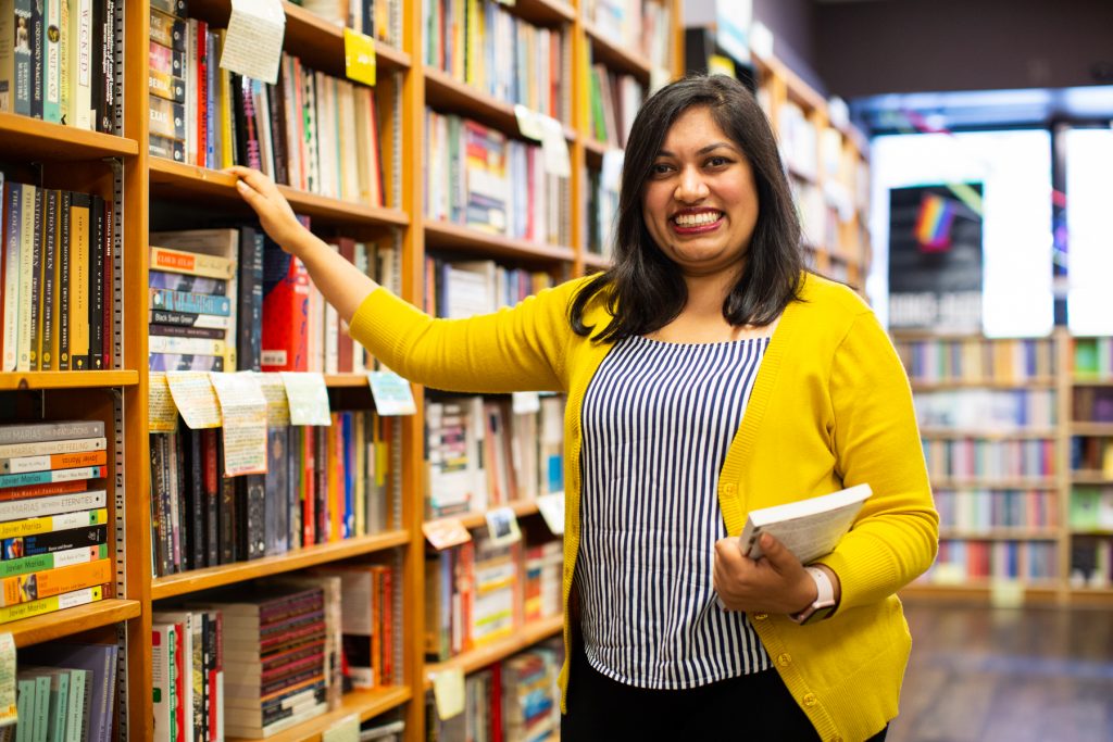 Image: Sharanya Sharma. Sharma stands inside a bookstore, smiling at the camera, with one hand holding a book and the other hand resting on a shelf. Sharma wears a marigold cardigan open over a black and white striped shirt, with black pants. The bookstore is bright with daylight. A long, tall set of shelves occupies most of the background. Photo by Kristie Kahns Photography.
