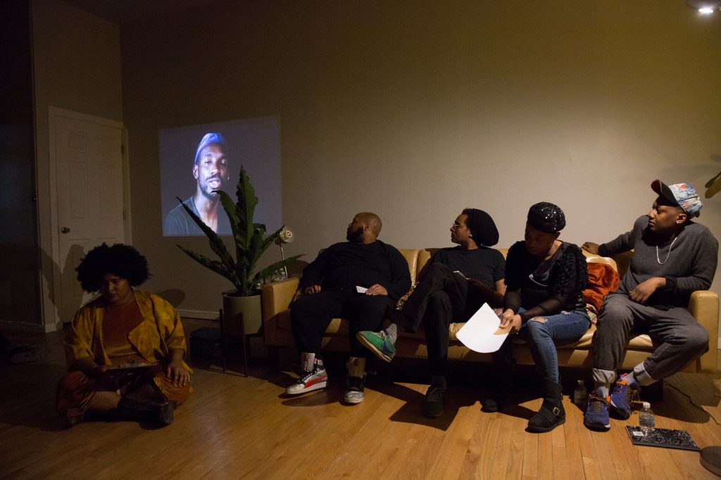 Image: In-Session, “Eclipsing: Migration, Movements and Desire,” at Threewalls in March 2018, presented by Amina Ross with J’Sun Howard, Khadijah Ksyia, Jared Brown, and A.J. McClenon, in response to the guiding work “Tongues Untied” by Marlon Riggs. In this shot, the artists sit in a row, with an image from “Tongues Untied” projected on the wall behind them in a low-lit room. Ross (left) sits on the wood floor and Howard, Brown, Ksyia, and McClenon (left to right) sit on a tan couch. Howard, Brown, and McClenon look toward the projected video and Ross and Ksyia look down. Ross wears a mustard-colored one-piece and jacket, Howard and Brown both wear all black, Ksyia wears a black top with blue jeans, and McClenon wears a grey top with grey jeans. Brown, Ksyia, and McClenon all wear headwear, and Ksyia holds paper in hand. Photo by Milo Bosh. Courtesy of Threewalls.