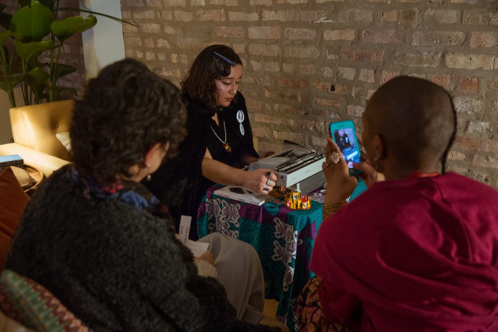  Image: In-Session, at Threewalls in November 2018, by Najee Searcy and collaborators, in response to the guiding work “Hush: Don’t Say Anything to God: Passionate Poems of Rumi” by Shahram Shiva. Melissa Castro Almandina sits at a small table that is covered in a colorful tablecloth, looking down at a typewriter while turning the knob to advance paper through it. Also on the table is a copy of Claudia Rankine’s book, “Citizen: An American Lyric,” and small sculptural objects that are primarily yellow, brown, and red. The artist wears a similar object as a pendant. The artist wears black clothes, as well as multi-colored rings, buttons, and hair clips. In the foreground, with their backs to the camera, sit two people, one using a phone to take a photo of the artist and the other holding slips of paper with text on them. Photo by Milo Bosh. Courtesy of Threewalls.