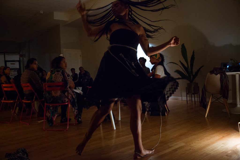 Image: In-Session, at Threewalls in November 2018, by Najee Searcy and collaborators, in response to the guiding work “Hush: Don’t Say Anything to God: Passionate Poems of Rumi” by Shahram Shiva. Ester Alegria of Zo//Ra performs in the foreground. Alegria is in mid-motion, as if spinning or twisting quickly, with arms out and one foot off the ground, and long braids and the bottom of a black dress flying outwards. The artist wears a black strapless dress, with a length of silver beads tied around its bodice and extending onto the ground, under and around the performer’s left foot. In the background of the dim room, audience members sit in a circle, most of them turned toward the performer. Photo by Milo Bosh. Courtesy of Threewalls.