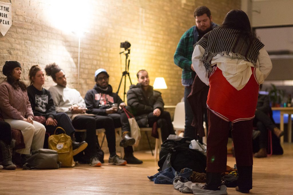 Image: In-Session, at Threewalls in February 2018, by Jose Luis Benavides and collaborators, in response to the guiding work “Mexican American Disambiguation” by José Olivarez. In the foreground, two performers stand, one facing the camera and one facing away from it, each wearing layers of varied clothes, including pants of varying lengths, multiple button-up shirts, and what appears to be a wrestling uniform. On the floor between them is a pile of still more clothes. In the background, several audience members watch from their chairs. Some audience members wear coats and other cold-weather clothes. Photo by Milo Bosh. Courtesy of Threewalls.
