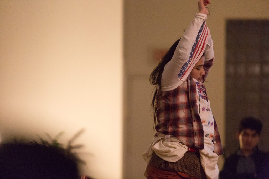 Image: In-Session, at Threewalls in February 2018, by Jose Luis Benavides and collaborators, in response to the guiding work “Mexican American Disambiguation” by José Olivarez. In this medium shot, a participant is in the act of dressing or undressing, pulling a white shirt with a U.S. American flag pattern over their head. The participant’s arms are in the air, entangled in the shirt, and underneath it the participant wears several tops and bottoms. One audience member is fuzzy in the background with, possibly, one in the foreground. Photo by Milo Bosh. Courtesy of Threewalls.