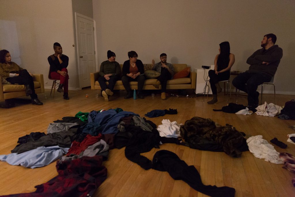 Image: In-Session, at Threewalls in February 2018, by Jose Luis Benavides and collaborators, in response to the guiding work “Mexican American Disambiguation” by José Olivarez. In the background, seven people sit on chairs or a tan couch, all looking towards the center, at Benavides, who is speaking. In the center of the low-lit room — in the foreground and around the image — is a sprawling pile of clothes. Photo by Milo Bosh. Courtesy of Threewalls.