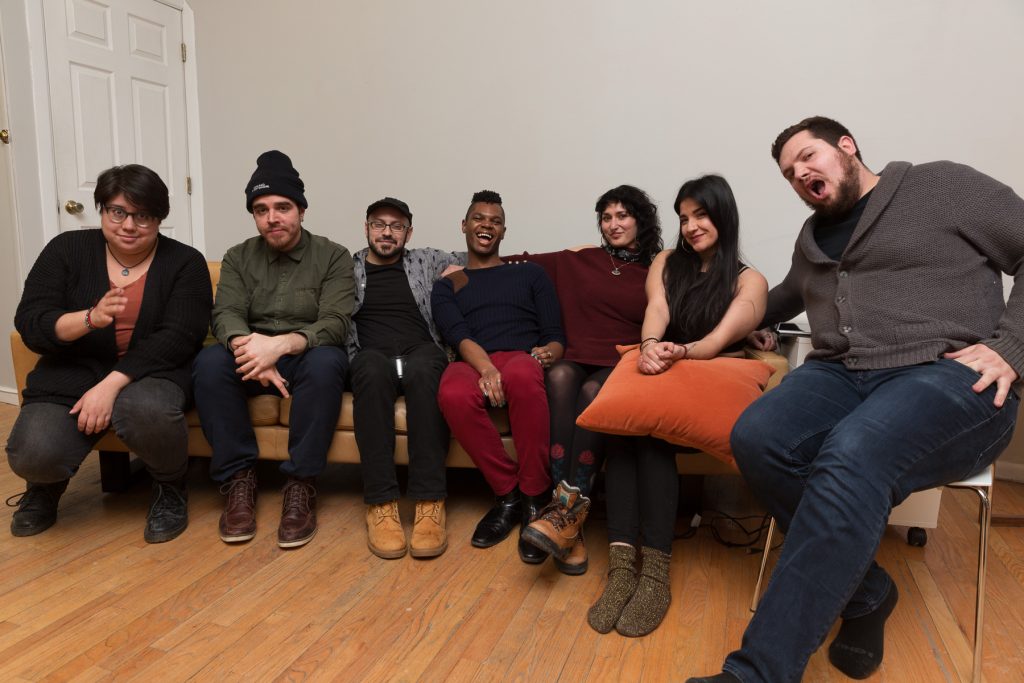 Image: In-Session, at Threewalls in February 2018, by Jose Luis Benavides and collaborators, in response to the guiding work “Mexican American Disambiguation” by José Olivarez. After the performance and discussion, Regina Martinez (third from right) and six others (performers) sit together on or near a tan couch, smiling or making faces at the camera. Martinez wears a maroon top with dark leggings and boots. Photo by Milo Bosh. Courtesy of Threewalls.
