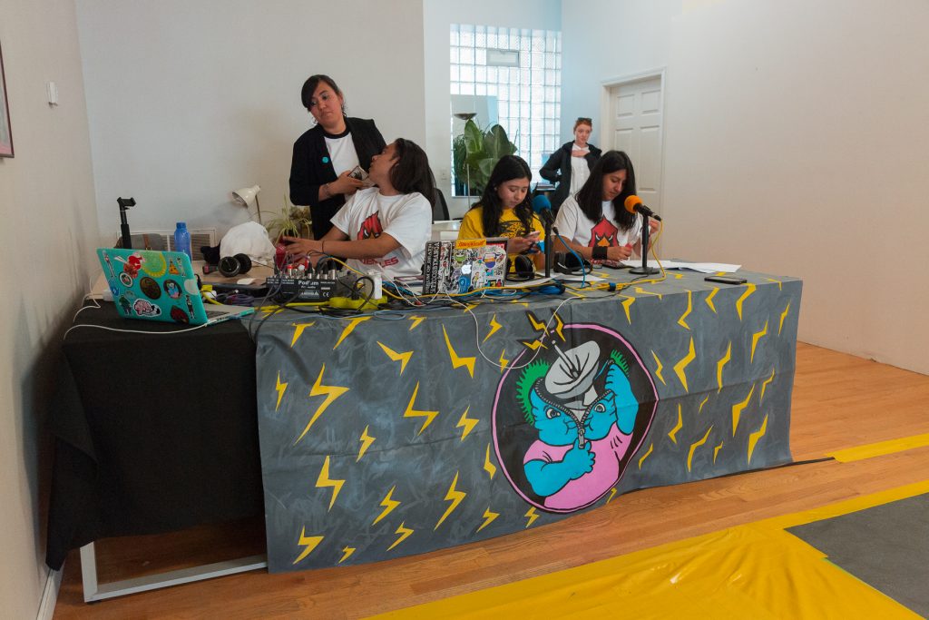 Image: In-Session, at Threewalls in June 2018, by El Cardenal De Aztlán and collaborators, in response to the guiding work “Borderlands/La Frontera” by Gloria Anzaldúa. Three artists sit behind a long table in the foreground and two stand behind them, one near, one farther back. On the table is a detailed dark grey covering (illustrated with yellow z-shapes like lightning bolts, as well as a circular inset showing a blue character unzipping its head open to reveal an active broadcast dish) and on that sit two microphones, two laptops, a soundboard, and other pieces of technology. The two artists seated behind the microphones each wear a t-shirt that matches the off-camera performers (a yellow t-shirt with a jaguar or a white t-shirt with a cardinal) and look down. The third artist wears the cardinal t-shirt and sits behind a soundboard, twisting back to talk to a person behind them. A yellow section of floor is visible in the lower-right corner of the frame. Photo by Milo Bosh. Courtesy of Threewalls.