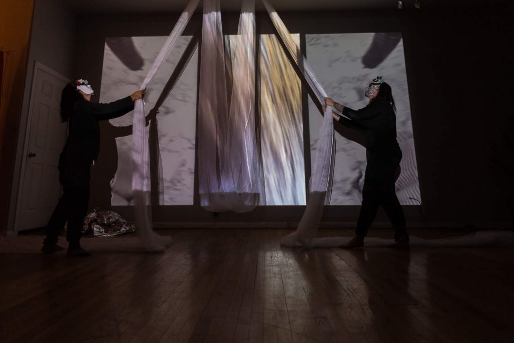 Image: In-Session, at Threewalls in March 2019, by Maya Mackrandilal with collaborators Udita Upadhyaya and Enid Muñoz and original performance for video by Bhanu Kapil, in response to the guiding work “Schizophrene” by Bhanu Kapil. In this wide shot, Muñoz (left) and Mackrandilal (right) perform. Both stand near a far wall, wearing black clothes and white masks with multi-colored decorations, looking up at a very long, diaphanous white cloth that is attached to the ceiling above them in two places with its middle portion hanging low between them. Each performer holds one part of the white cloth, with each end of its remainder snaking off-camera to the left and right behind each performer. A wall-sized video, showing a textured white scene, is projected over the performers and the cloth. Balled up in one corner of the floor is a silver reflective cloth. Photo by Milo Bosh. Courtesy of Threewalls.