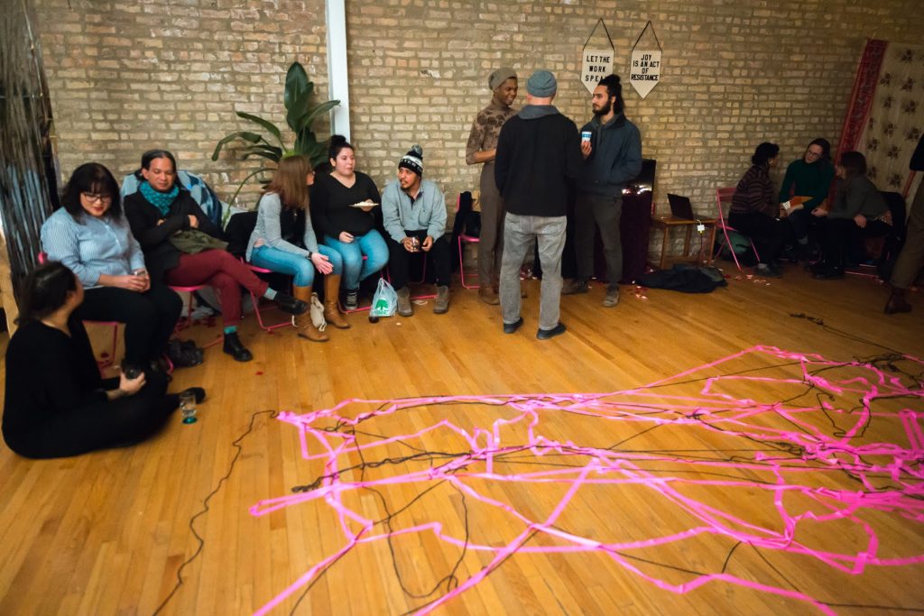 Image: In-Session, at Threewalls in March 2019, by Maya Mackrandilal with collaborators Udita Upadhyaya and Enid Muñoz and original performance for video by Bhanu Kapil, in response to the guiding work “Schizophrene” by Bhanu Kapil. In the background of this shot, trios of audience members and performers sit or stand around the room, chatting after the performance and discussion. Remnants of the performance are in the foreground — a layered web of pink fluorescent ribbon and black string on the wood floor, reflecting where many audience members stood during one part of the performance. Many audience members wear hats, sweaters, or other cold-weather clothes. Photo by Milo Bosh. Courtesy of Threewalls.