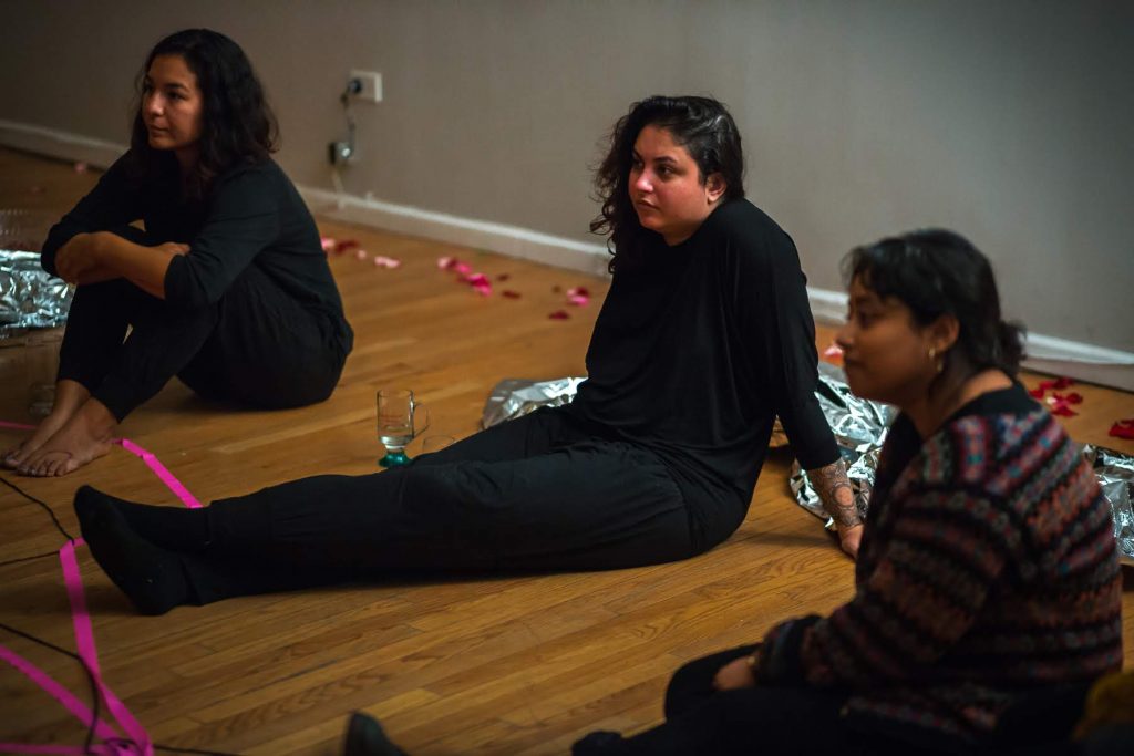 Image: In-Session, at Threewalls in March 2019, by Maya Mackrandilal with collaborators Udita Upadhyaya and Enid Muñoz and original performance for video by Bhanu Kapil, in response to the guiding work “Schizophrene” by Bhanu Kapil. Muñoz (left), Mackrandilal (center), and Upadhyaya (right) sit on the wood floor during the discussion portion of the event. They sit in oblique profile to the camera, looking off-camera toward the audience in the low-lit room. Muñoz and Mackrandilal both wear all black, and Upadhyaya wears a patterned sweater with dark bottoms. A length of fluorescent pink ribbon sits in the lower-left corner of the frame. Photo by Milo Bosh. Courtesy of Threewalls.