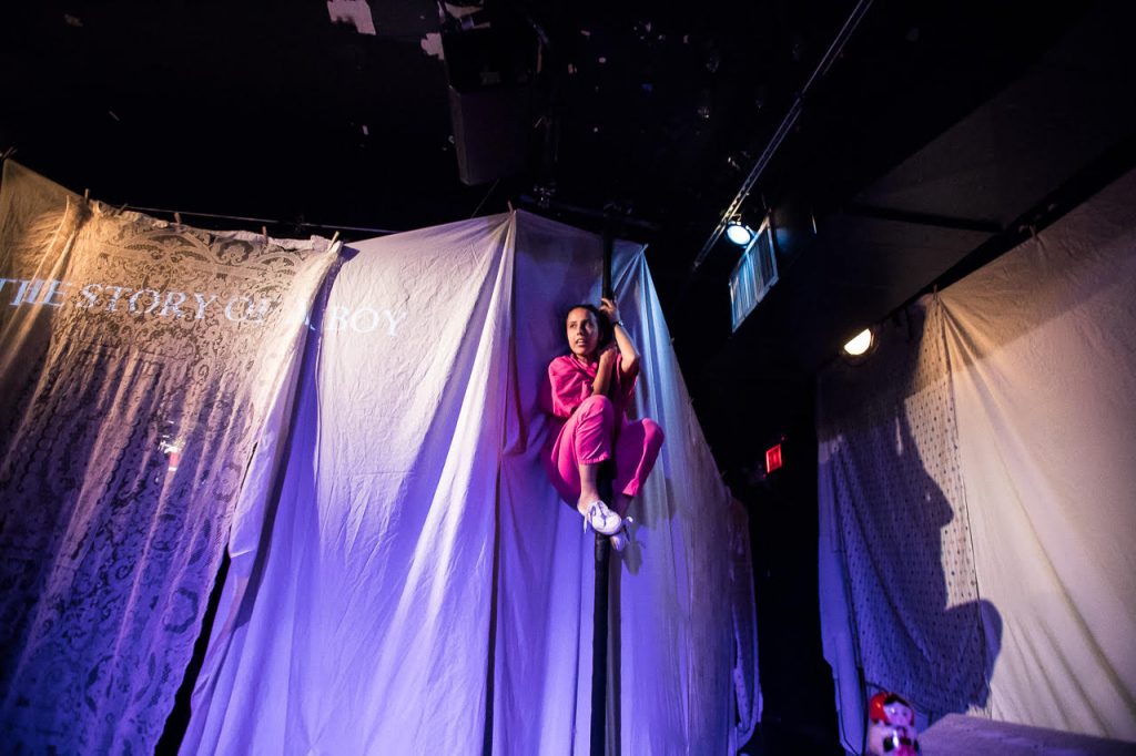 Image: Center, Ida holding herself in a crouch position on a pole in front of a wall of curtains. To the left at level with Ida's head, projected across a taffeta sheet and a bedsheet are the words "THE STORY OF A BOY." To the right, the sheets taper off into a hall and at the top, a red exit sign. Further right, Ida's shadow on the wall. Below that on top of a box, a Russian nesting doll. Photo Courtesy of Brave Lux, Inc.