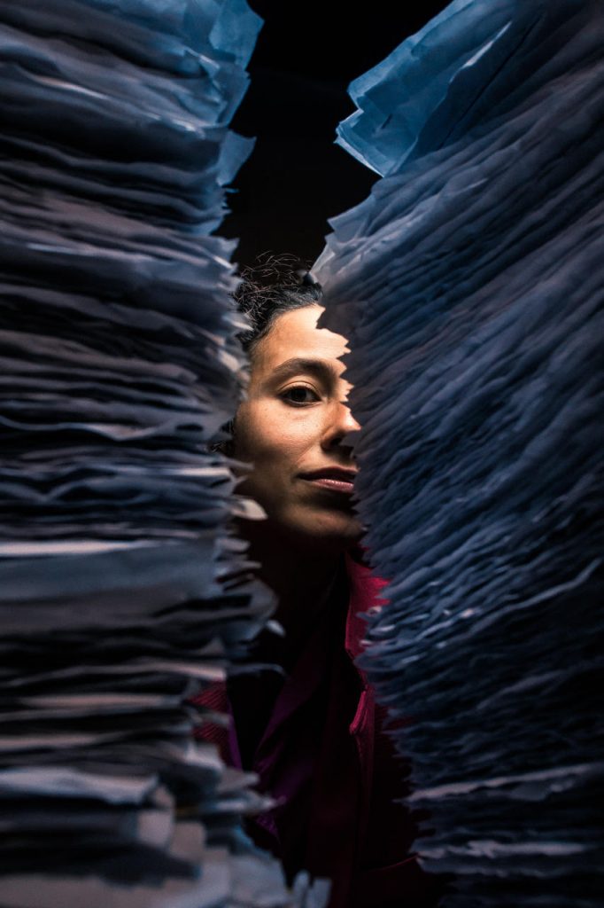 Image: Two stacks of wrinkled, fringed, blue-tinted papers stand close to the camera, blurred. Between them, Ida Cuttler looks at the camera with an almost-smile; the right side of her face is obscured by the stack of paper. Above and below her face is darkness. She wears a pink blazer. Photo courtesy of Brave Lux, Inc.