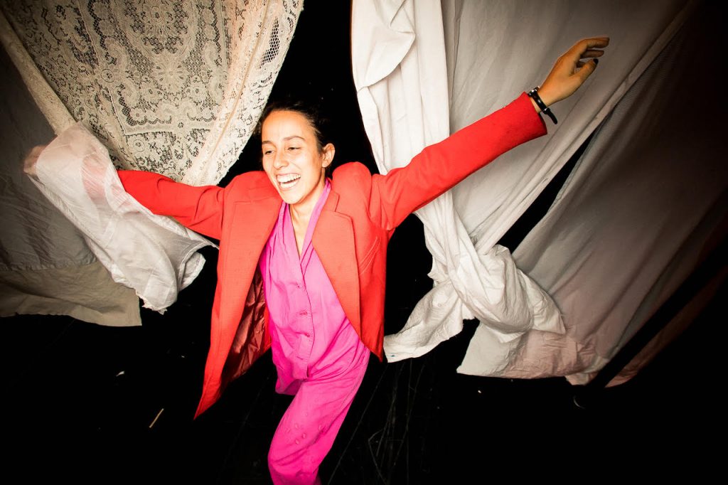 Image: Ida Cuttler, with a toothy smile and wearing a crimson blazer and pink pajamas, has her arms extended out.  Behind her is an opening between several twisting bedsheets hung to the right and a taffeta sheet hung to the left. Her left hand has caught some of a transparent sheet. Photo courtesy of Brave Lux, Inc.