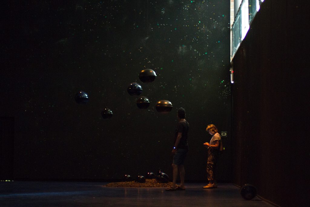 Image: Viewers interacting with "Dark Matter: Celestial Objects as Messengers of Love in These Troubled Times" by Folayemi Wilson at the Hyde Park Art Center. The installation has made the space very dark with hanging orbs towards the middle of the frame. Photo by Michael Sullivan.