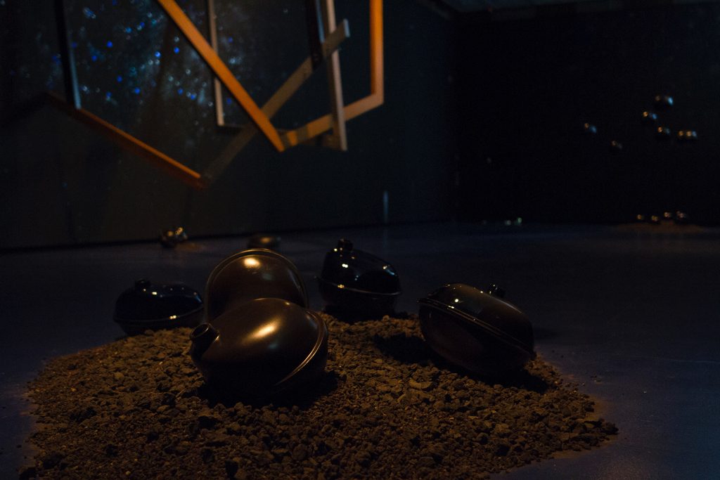 Image: 'Fallen objects' from outer space within the installation "Dark Matter: Celestial Objects as Messengers of Love in These Troubled Times" by Folayemi Wilson at the Hyde Park Art Center. Dirt lay on the floor with sculptural objects on and suspended above. Photo by Michael Sullivan.