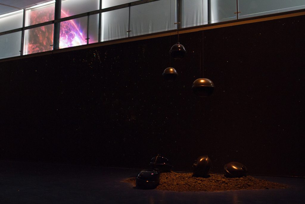 Image: Installation view of "Dark Matter: Celestial Objects as Messengers of Love in These Troubled Times" by Folayemi Wilson at the Hyde Park Art Center. Objects are hanging in the air and 'crash landing' onto a pile of earth. Photo by Michael Sullivan.