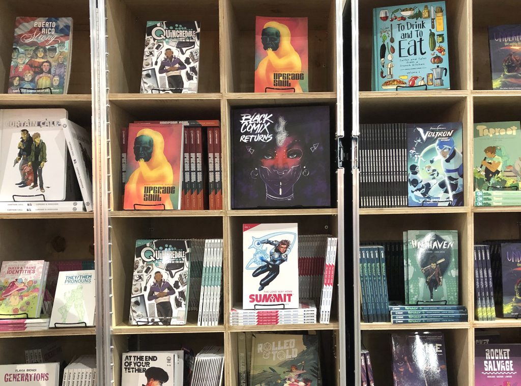 Image: The Black Comix Returns book sits on a wood bookshelf surrounded by other books, mostly graphic novels, at San Diego Comic Con 2019. The Black Comix Returns cover is various shades of purple and features a drawing of a woman’s face, with pink across the bridge of her nose and her cheeks. The artwork on the cover is by Ashley A. Woods. Photo by Jessica Hammie