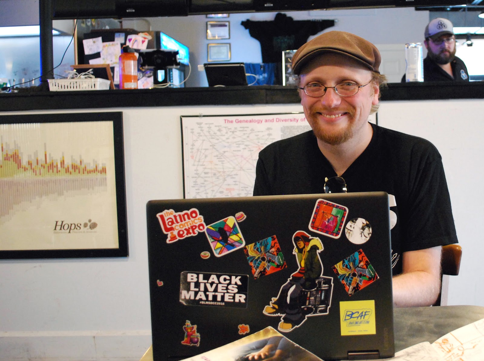 Image: Damian Duffy is seated at a table in a brewery. He is smiling and looking at the camera. In front of him is a laptop computer, covered in stickers. Photo by Jessica Hammie.