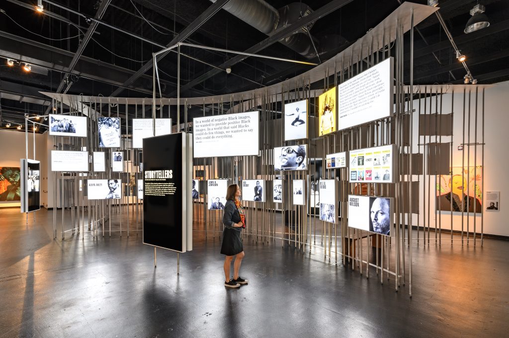Image: Installation view of Men of Change. The installation made of metal poles is in the middle of the room. Light boxes of various sizes featuring photos and text are hung on the metal poles. A large display box in front of the installation features a poster that reads STORYTELLERS. Along the walls of the room are paintings and other artworks, most of which are obscured by the metal pole installation in the middle of the room. A woman stands in front of the light box installation, looking at the installation. Photo by Phil Armstrong. Photo courtesy of the Smithsonian Institution Traveling Exhibition Service. 