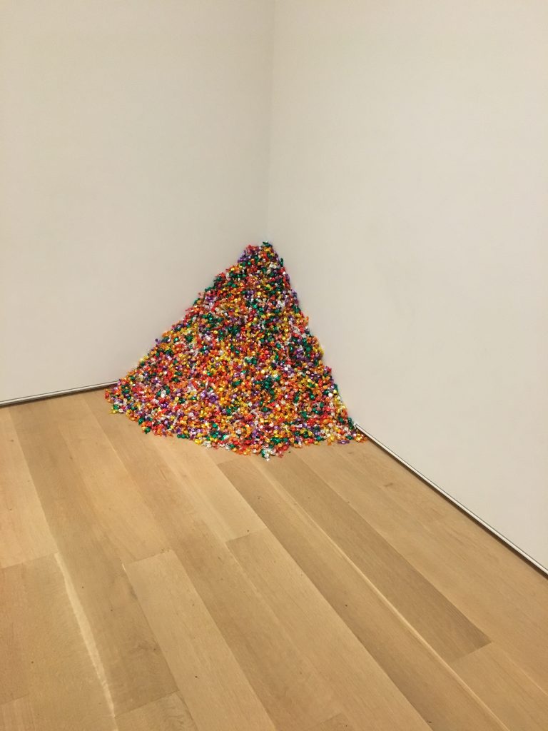 Image: Félix González-Torres' Untitled (Portrait of Ross): a spill of colorful wrapped candies piled in the corner of a gallery space. Red, green, yellow, and purple candies make a 175 pound mountain, from which viewers are welcome to take a piece home with them or eat. The museu takes responsibility for replenishing the pile to maintain the dictated weight. Image by the author. 