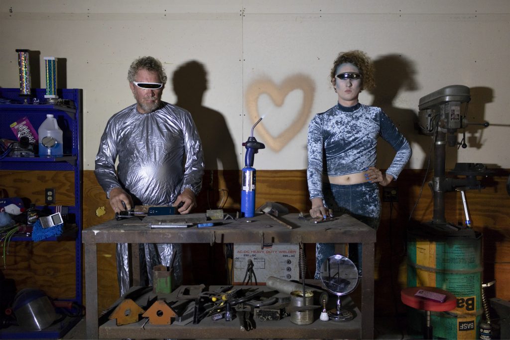 Image: FarmBot 3000 and FemmeBot 6000, 2018 by Nadia Stiegman. Two Cyborgs are shown in a tool shed standing side by side with various tools and toys around them. A heart is spray painted to the back wall. Image courtesy of the artist.