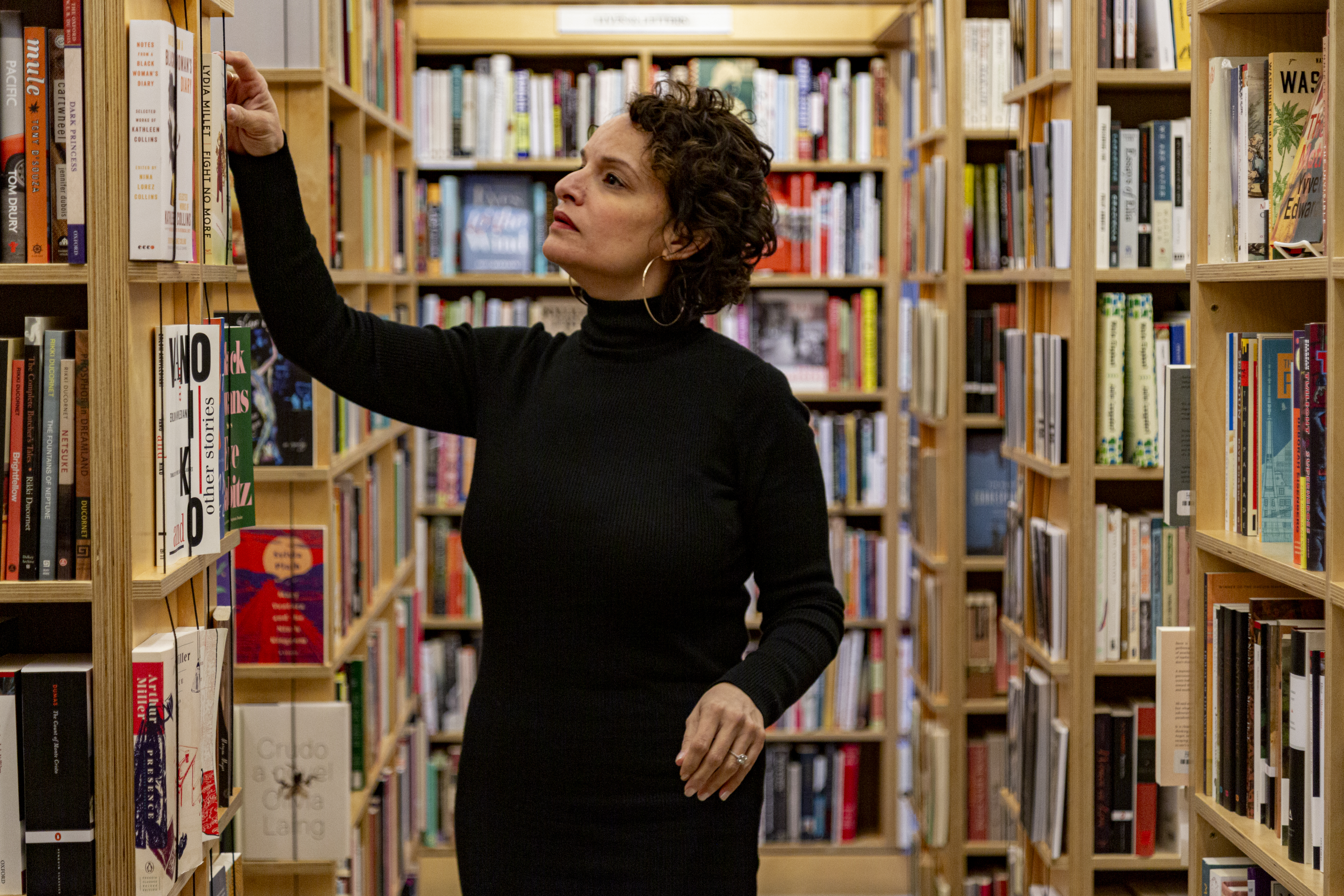Image: Mustafah stands inside a bookstore, reaching up to pull a book off a shelf. She wears a long-sleeved black turtleneck dress and large hoop earrings. Bookshelves full of books fill the space behind and around her. Photo by Mark Blanchard.