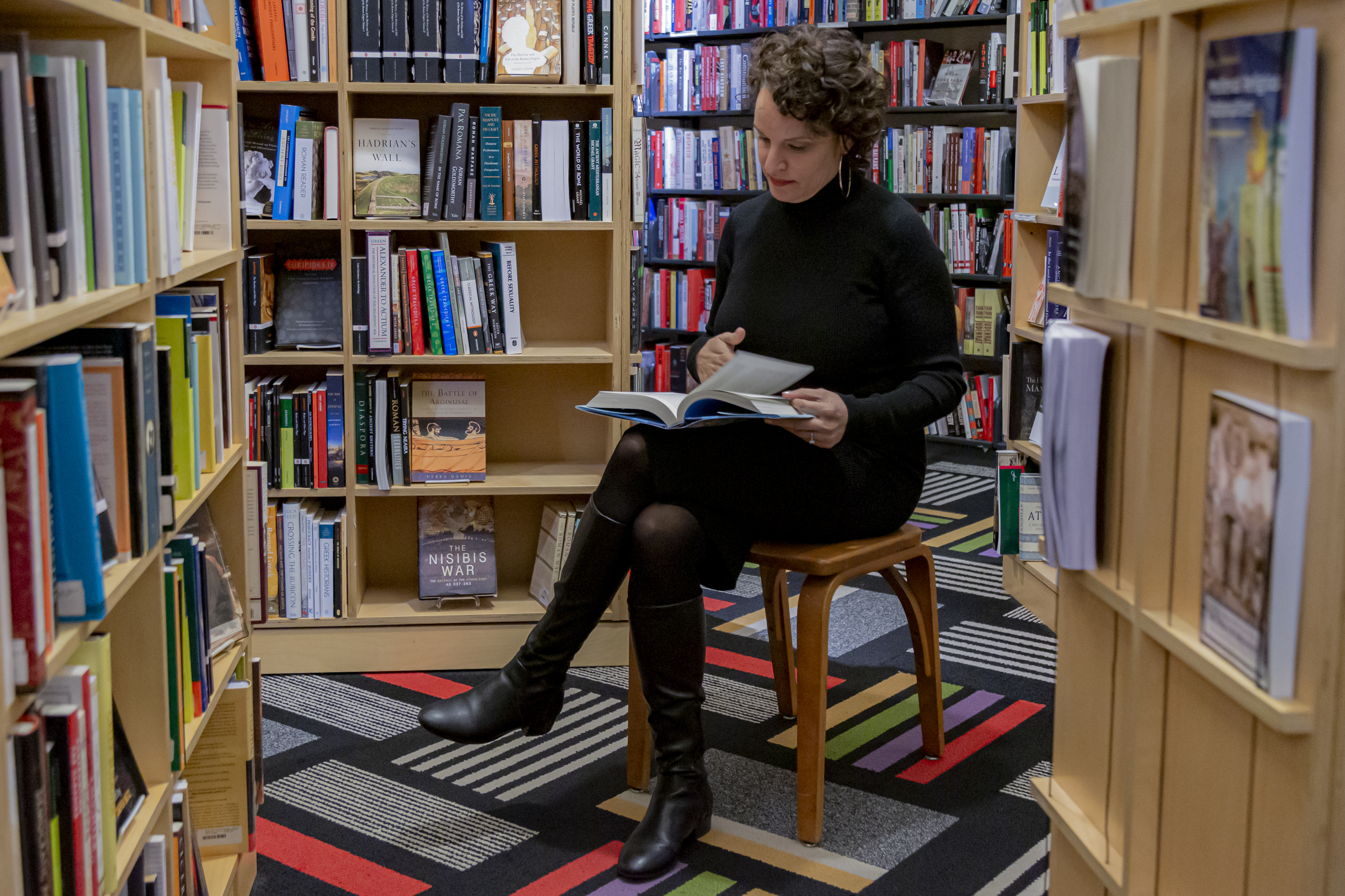 Image: Mustafah sits in a wooden chair inside a bookstore, browsing through an open book in her lap. She wears a long-sleeved black turtleneck dress, black tights, black boots, and large hoop earrings. Bookshelves full of books are behind and around her, positioned at various angles. The carpet’s pattern is a grid of thin and thick stripes in black and white and bright colors. Photo by Mark Blanchard.