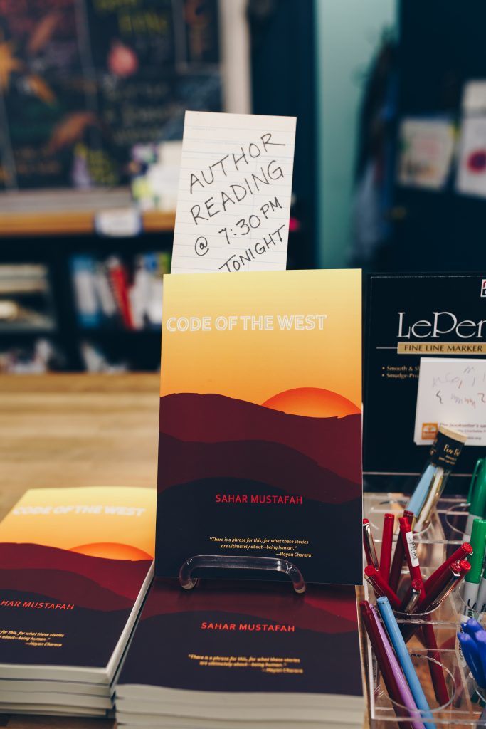 Image: Mustafah’s short story collection, “Code of the West” (Willow Books, 2017). One copy of the book is displayed upright atop several other copies, which lay flat on a bookstore countertop. A hand-written paper reads, “Author reading @ 7:30pm tonight,” in all capital letters. The book cover’s illustration shows a sliver of orange sun behind reddish and dark brown hills, with a yellow sky above. Photo by Michelle Strahan.