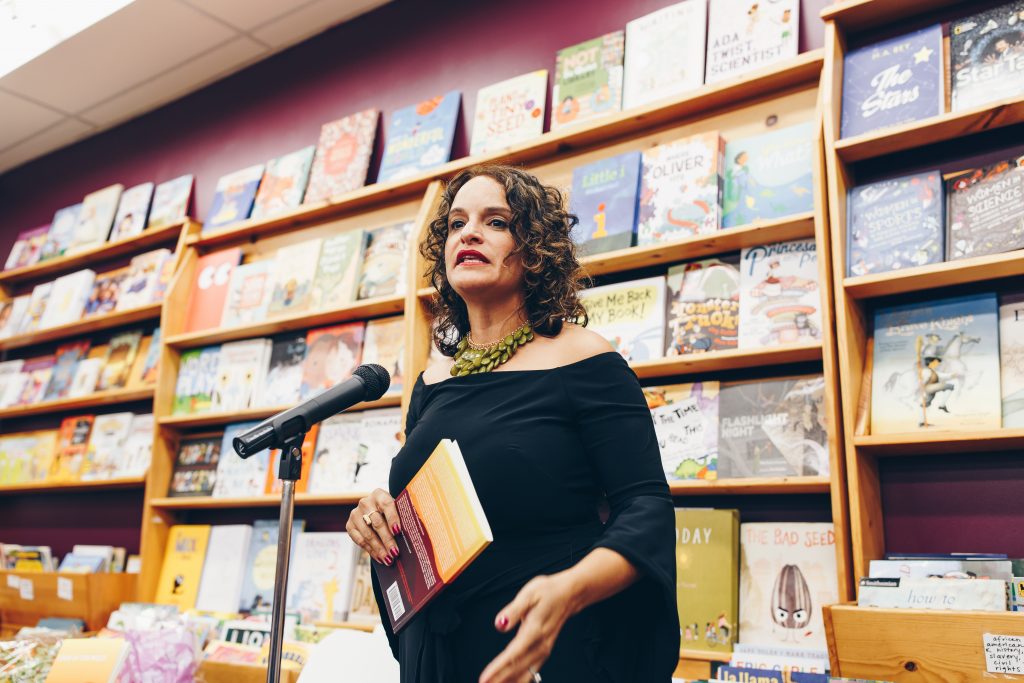 Image: Mustafah reading at the book launch event for her short story collection, “Code of the West,” at Women & Children First Bookstore in 2017. Mustafah stands, speaking into a microphone, a wall of picture books behind her. In her hand, she holds a copy of “Code of the West.” Mustafah wears a dramatic green necklace and an off-the-shoulder black shirt with three-quarter-length sleeves that drape open at the hem. Photo by Michelle Strahan.