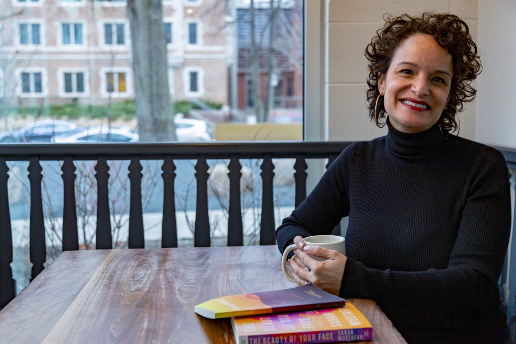 Image: Sahar Mustafah. Mustafah sits at a wooden table, holding a coffee cup and smiling at the camera. In the foreground are two books, her short story collection and her debut novel; their covers are mostly a mix of yellows, oranges, pinks, and browns. Mustafah wears a long-sleeved black turtleneck and large hoop earrings. Behind her is a Hyde Park street, and natural light comes through the window. Photo by Mark Blanchard.