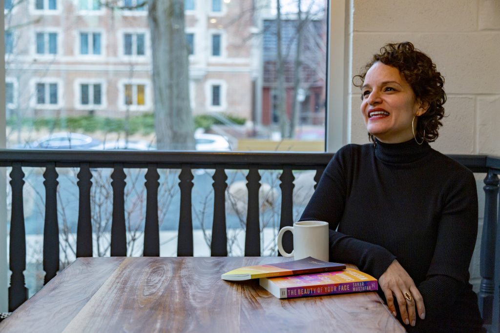 Image: Mustafah sits at a wooden table, looking off-camera and laughing. In the foreground are a coffee cup and two books, her short story collection and her debut novel; the books’ covers are a mix of mostly yellows, oranges, pinks, and browns. Mustafah wears a long-sleeved black turtleneck and large hoop earrings. Behind her is a Hyde Park street, and natural light comes through the window. Photo by Mark Blanchard.