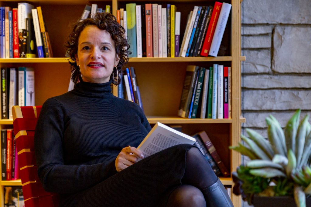 Image: Sahar Mustafah sits in a red chair inside the Seminary Co-op Bookstore, holding a closed book in her lap and smiling at the camera. She wears a long-sleeved black turtleneck dress, black tights, black boots, and large hoop earrings. Books are scattered on the bookshelves behind her. A plant sits in the foreground and a stone wall is partially visible in the background. Photo by Mark Blanchard.