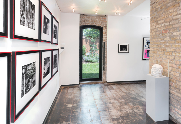  Image: Installation shot of Iceberg Projects exhibition “David Wojnarowicz: Flesh of My Flesh,” June 23 – August 5, 2018. On the left wall, a series of images are hung in rows. A sculpture of a head is encased in glass on the right way. Directly in front of the viewer, an outdoor green space is visible through a glass door. Image courtesy of Iceberg Projects.  
