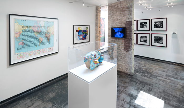 Image: Installation shot of Iceberg Projects exhibition “David Wojnarowicz: Flesh of My Flesh,” June 23 – August 5, 2018. In the middle of the room, a sculpture sits on a white pedestal, encased in glass. A world map with words overlaying the image is hanging on the wall to the left, and a video screen is displaying a video on the partial brick wall directly in front of the viewer. Image courtesy of Iceberg Projects.