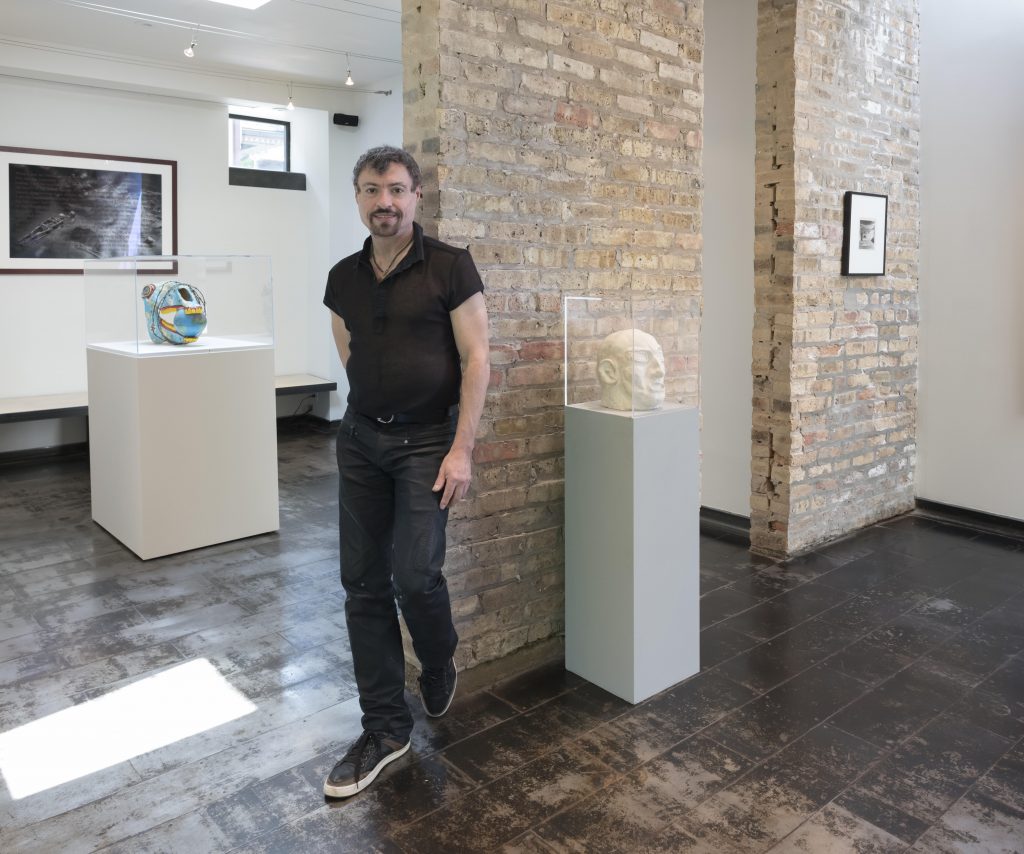 Image: Dr. Dan Berger standing against a partial brick wall in the Iceberg Projects exhibition space. He is wearing dark jeans and a black short-sleeve shirt. Image courtesy of the artist. 