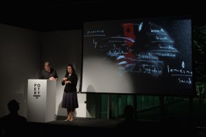 [IMAGE DESCRIPTION: Matt Bodett stands behind a white podium to the left, next to him is a woman, the American Sign Language interpreter, she is making a sign with her hands, they are both in a spotlight. Behind them is a projection screen showing a still from a video that features primarily white chalk-like marks and fragmented words on a black background, there is a swath of red on the screen as well.] Photo by Ryan Thiel.