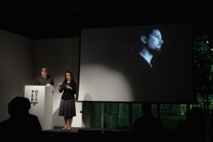[IMAGE DESCRIPTION: Matt Bodett stands behind a white podium to the left, next to him is a woman, the American Sign Language interpreter, she is making a sign with her hands, they are both in a spotlight. Behind them is a projection screen showing a still from a video of the artist. We see Bodett, in black and white on a black background, from a profile angle and from the waist up.] Photo by Ryan Thiel.