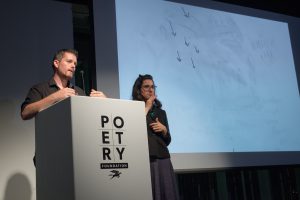 [IMAGE DESCRIPTION: Matt Bodett stands behind a white podium which bears the Poetry Foundation logo on the front, he caught mid-speech. To his right is a woman, the American Sign Language interpreter, she is making a sign with her hands. To their right is a projection screen which shows a primarily white screen with small pencil-like marks on it, the marks look like six small arrows pointing downward.] Photo by Ryan Thiel.