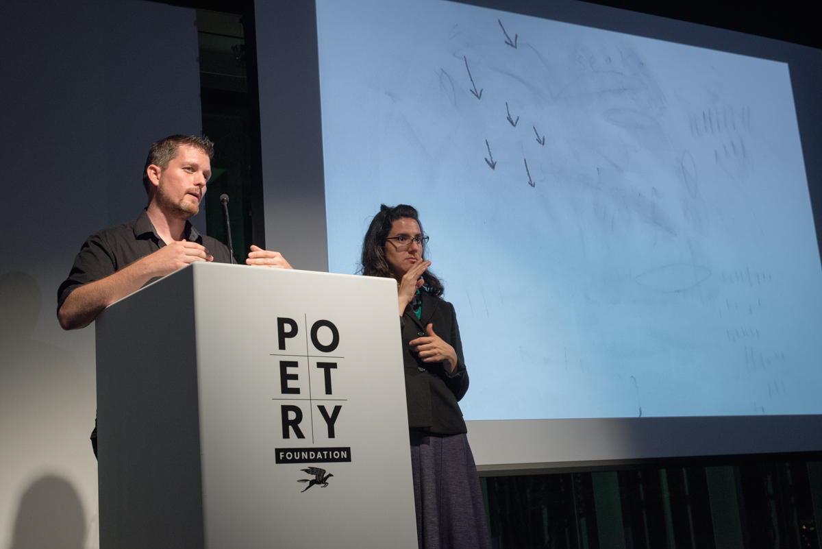 [IMAGE DESCRIPTION: Matt Bodett stands behind a white podium which bears the Poetry Foundation logo on the front, he caught mid-speech. To his right is a woman, the American Sign Language interpreter, she is making a sign with her hands. To their right is a projection screen which shows a primarily white screen with small pencil-like marks on it, the marks look like six small arrows pointing downward.] Photo by Ryan Thiel.
